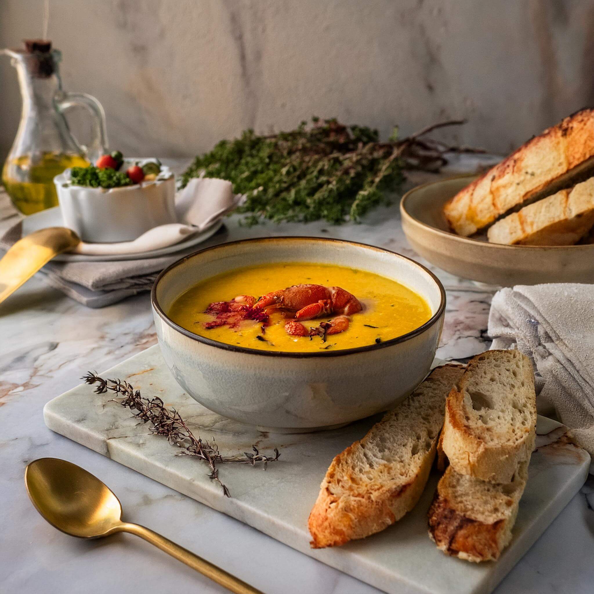 Final presentation of lobster bisque with bread, herbs, and tableware on a white marble countertop.