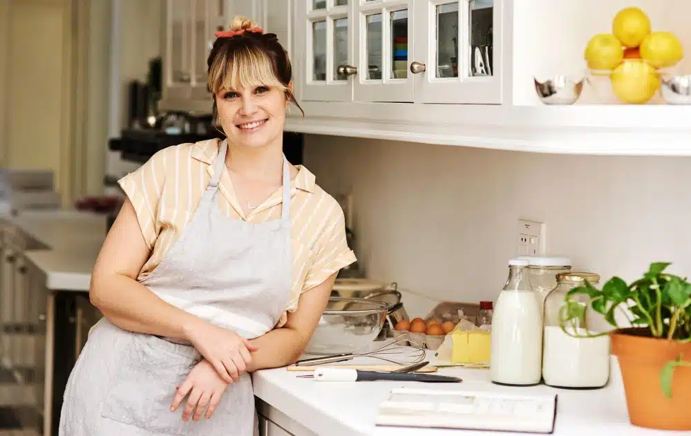 A woman in a light apron leans on a kitchen counter with baking ingredients and a recipe book.