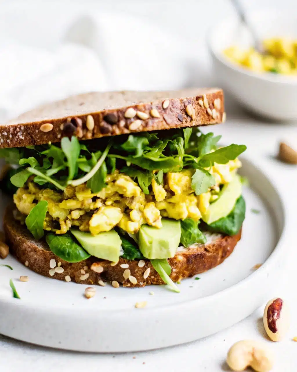 Curry chicken salad sandwich on whole grain bread with avocado slices and fresh arugula on a white plate.