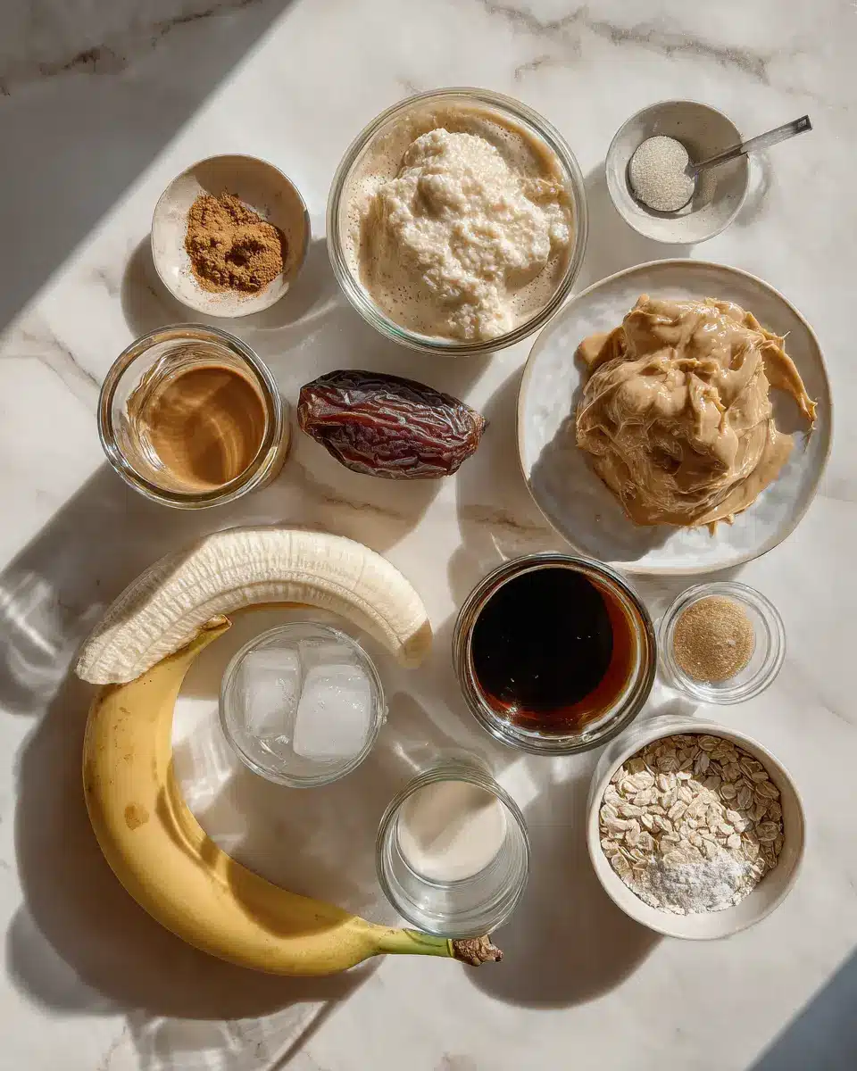 Flat-lay of 1 frozen banana, espresso shot, cashew butter, almond or oat milk, vanilla extract, Medjool date or maple syrup, vanilla protein powder, and ice cubes on a white marble kitchen counter in warm natural light.